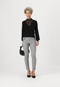 Black sweater with a lace panel, paired with gray plaid trousers featuring zip details. Model holds a small black handbag and wears black heels.