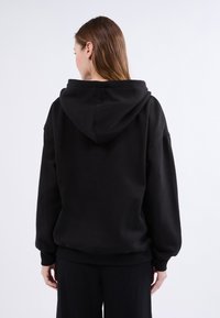 Sudadera con capucha negra hecha de tela suave. Presenta un corte holgado, mangas largas y puños acanalados, con una gran capucha en la parte posterior.