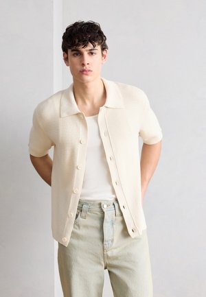 Young man with curly hair wearing a white knit short-sleeve cardigan over a white tank top and light beige jeans, standing against a plain background.