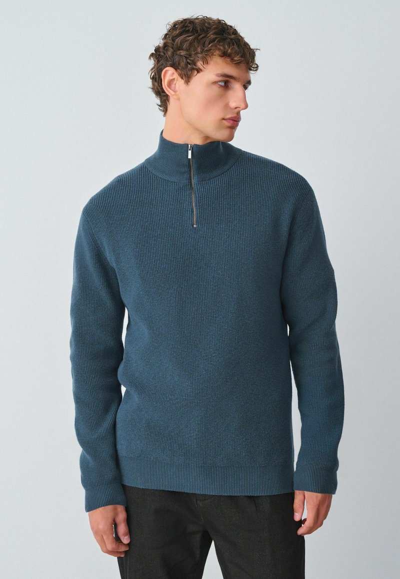 Maglione lavorato a maglia color teal con colletto alto e chiusura a mezza zip. Tessuto testurizzato con polsini e orlo a costine. Indossato sopra pantaloni scuri.