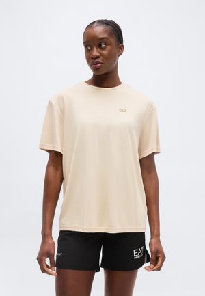 Kvinde iført en beige oversized T-shirt og sorte shorts, stående mod en ensfarvet lys baggrund, kiggende til siden.