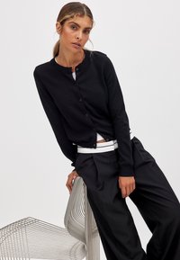Schwarze gestrickte Strickjacke mit rundem Halsausschnitt und Knopfverschluss. Kombiniert mit lockeren schwarzen Hosen, sitzt auf einem Drahtstuhl mit elegantem Design.
