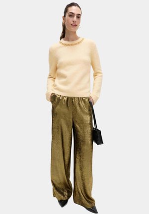 Suéter de punto amarillo claro con un detalle de cadena en el escote, combinado con pantalones anchos de color dorado brillante y zapatos negros de punta afilada.