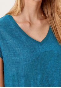 Top de lino teal de manga corta con cuello en V y diseño de hojas bordadas con textura en la parte frontal. Tejido ligero y suave.