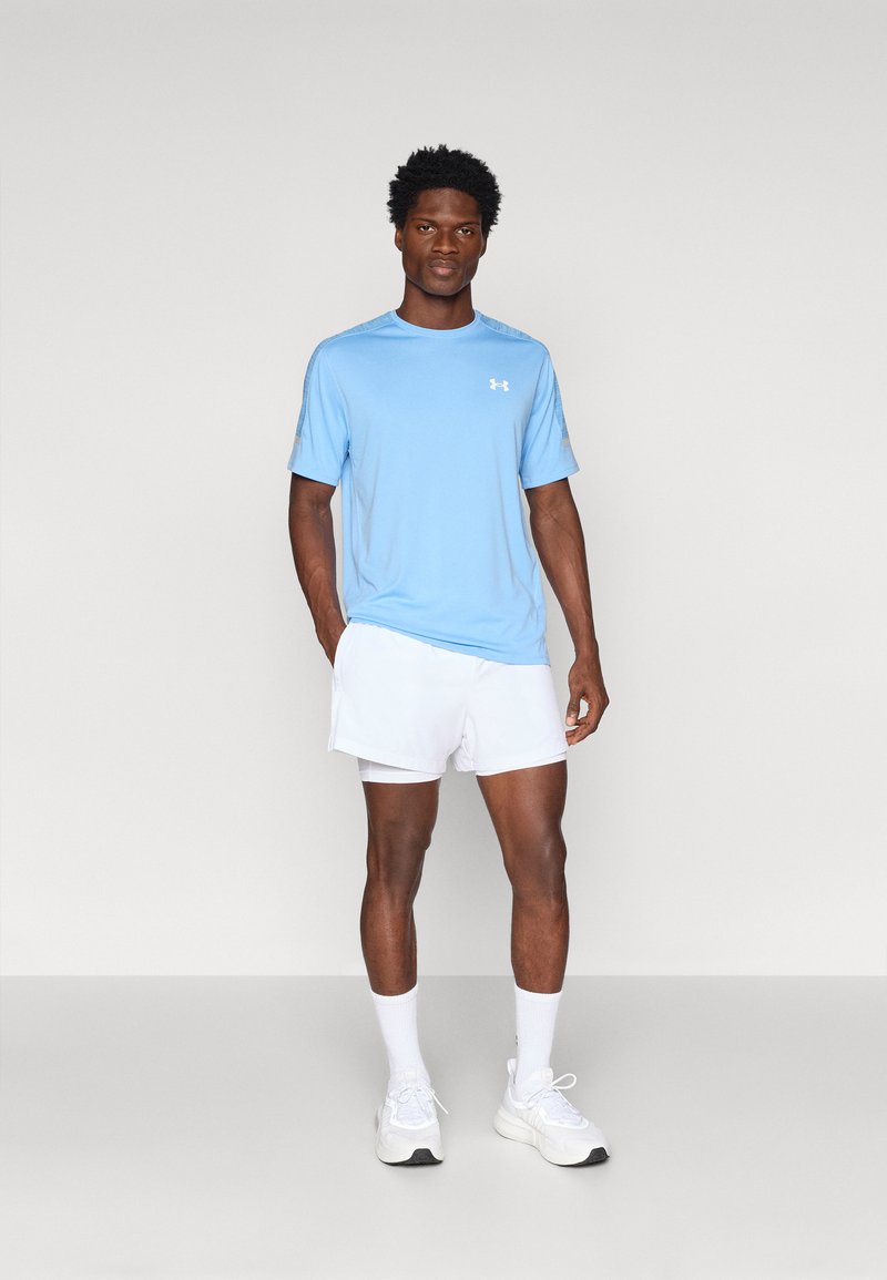 Maglietta atletica azzurro chiaro con maniche corte e logo, abbinata a pantaloni corti bianchi. Il modello si trova su uno sfondo neutro, indossando calzini bianchi e sneakers.