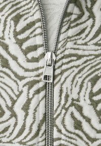 Nahaufnahme eines silbernen Reißverschlusses auf beige und olivgrün gemustertem Strickstoff mit abstraktem Blattdesign.