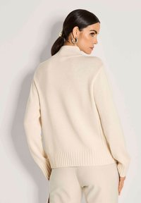 Frau mit dunklem Haar im Pferdeschwanz steht seitlich, trägt einen cremefarbenen, gerippten Rollkragenpullover und hellfarbene Hose vor einem einfarbigen Hintergrund.