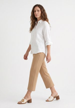 Kvinde med langt bølget hår går iført en hvid skjorte med knapper, beige korte bukser og cremefarvede højhælede sandals med cirkulære pynt.