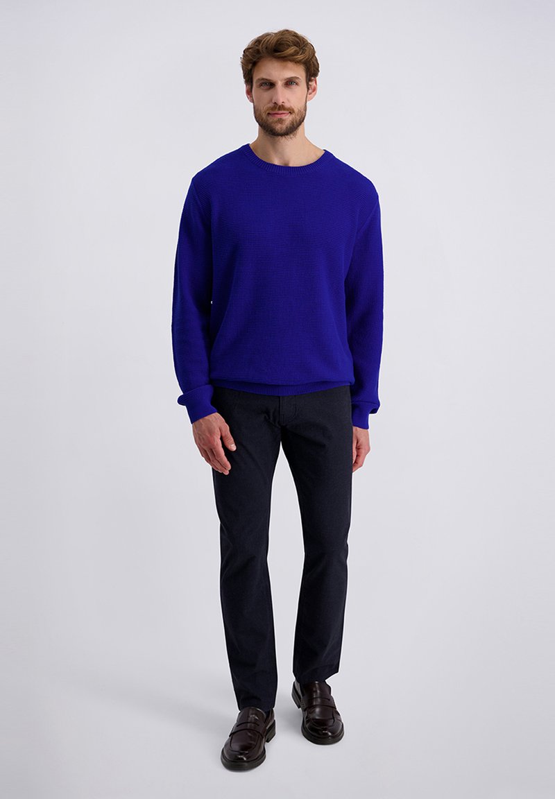 Homme debout portant un pull à manches longues bleu vif, un pantalon noir et des mocassins marron foncé contre un fond clair neutre.