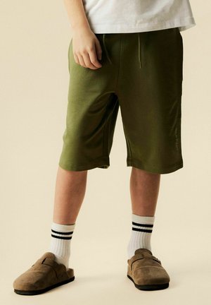 Dziecko ubrane w oliwkowo-zielone szorty do kolan, białe skarpetki z czarnymi paskami i brązowe buty wsuwane na beżowym tle.