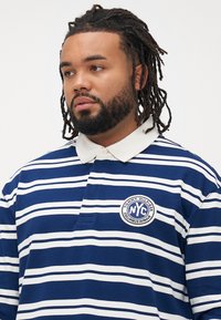 Mann med dreads og skjegg som har på seg en marineblå og hvit stripete polo med hvit krage og et rundt NYC-logo-patch på brystet.