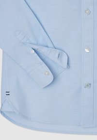 Jasnoniebieska koszula z długim rękawem o fakturowanym wykończeniu, z przezroczystymi guzikami, regulowanymi mankietami i kontrastującymi czarnymi paskami u dołu.