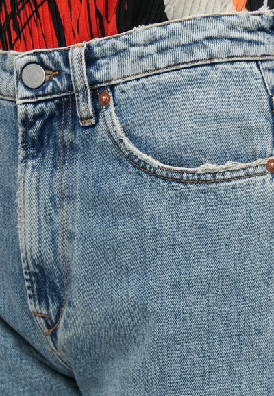 Φωτεινό γαλάζιο τζιν παντελόνι με ξεφτισμένο τελείωμα, χαλκή κουμπί και ορατή ραφή. Το ύφασμα εμφανίζει ξεθωριασμένη υφή με διακριτικές παραλλαγές.