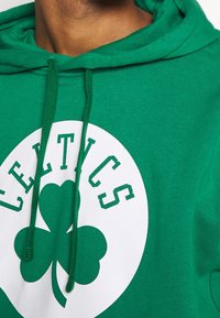 Haut à capuche en coton vert avec un grand logo circulaire blanc portant le mot "CELTICS" et un motif de trèfle à quatre feuilles au centre.