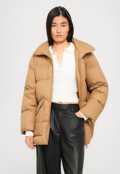 Frau in einer sandfarbenen Steppjacke über einem weißen Stricktop und schwarzen Lederhosen, steht mit einer Hand in der Tasche vor einem schlichten Hintergrund.