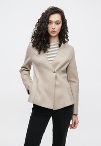 Beige jack van een wolmix met een nauwsluitend silhouet, sluiting met één knoop en schuine naden. Gedragen over een zwart-wit gestreept shirt.