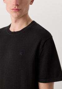 Zwarte katoenen T-shirt met een ronde hals, korte mouwen en een subtiel geborduurd logo op de linkerkant van de borst. Gladde textuur, minimalistisch ontwerp.