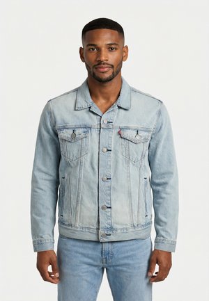 Uomo in piedi che guarda avanti, indossa una giacca di jeans azzurro chiaro e jeans abbinati, su sfondo bianco uniforme.