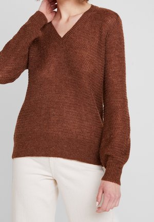Maglione in maglia marrone con scollo a V, maniche lunghe a palloncino e superficie texture. Abbinato a pantaloni chiari.