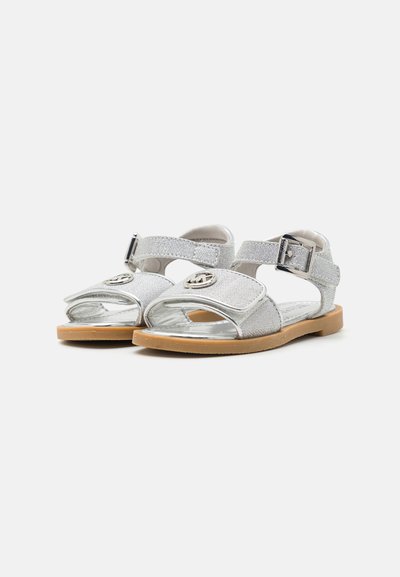 Sandales pour enfants argentées avec une finition brillante, bride ajustable, accent de logo circulaire et semelle en caoutchouc brun clair. Design ouvert sur l'avant.