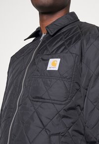 Černá prošívaná bunda se zapínáním na zip, límcem na patentky a náprsní kapsou s bílou čtvercovou etiketou s oranžovým logem.