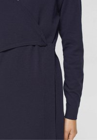 Mörkblå stickad klänning med omslagsdesign, långa ärmar och band i midjan för en figurnära silhuett. Slät textur med minimala detaljer.