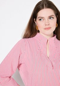 Blusa a righe rosse e bianche con colletto alla cinese, maniche lunghe e dettaglio con abbottonatura. Tessuto morbido con una finitura pulita.