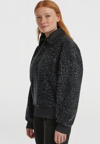 Vrouw met rood haar in een zwarte, getextureerde fleece jas met een hoge kraag en zwarte ribgebreide manchetten, poserend tegen een grijze achtergrond.