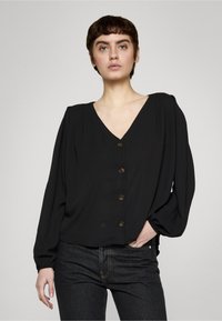 Blusa negra con escote en V, mangas largas y cierre de botones. Tejido suave, detalles plisados en los hombros y un corte holgado. Combinada con jeans oscuros.