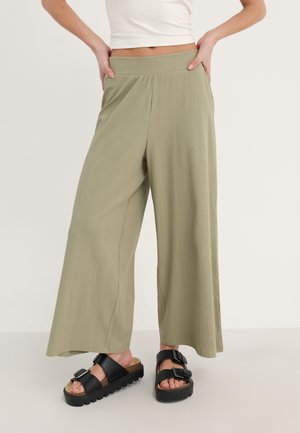 Pantalons larges, côtelés, de couleur olive avec une taille élastique, accompagnés de sandales noires à boucle sur un fond clair.
