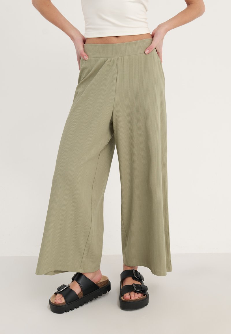 Wijduitlopende, ribbelige, olijfgroene broeken met een elastische tailleband, gecombineerd met zwarte sandalen met gespen op een lichte achtergrond.