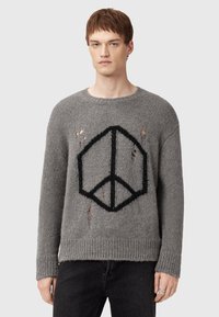 Giovane uomo che indossa un maglione grigio lavorato a maglia con simbolo della pace nero e buchi consumati, abbinato a pantaloni neri, in piedi contro uno sfondo bianco.
