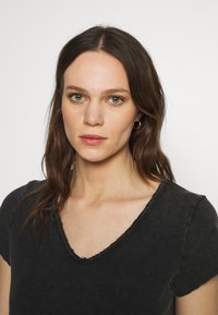 Melnā v-veida T-kreklā ar mīkstu tekstūru. Tam ir neapstrādātas, plīsušas malas apkaklē un piedurknēs. Vienkāršs, minimāls dizains.