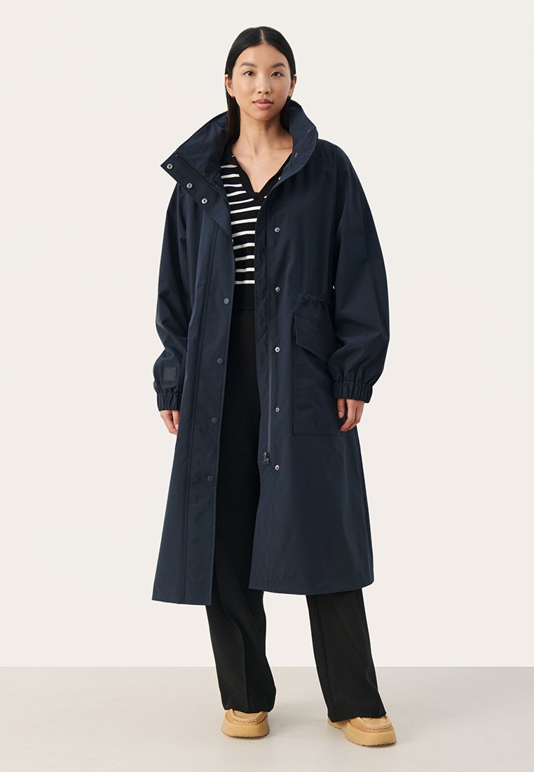 Cappotto lungo impermeabile blu navy con colletto alto, polsini elasticizzati e chiusura a pressione sul davanti. Presenta tasche laterali e una vestibilità comoda.