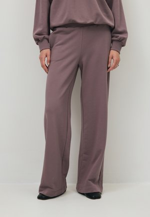 Persoon met losse mauve broek en bijpassende trui staand tegen een effen lichte muur, zwarte schoenen zichtbaar.