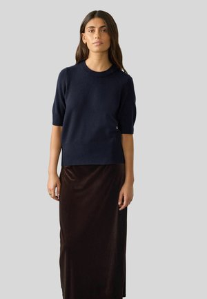 Námořnická modrá pletená halenka s krátkým rukávem a kulatým výstřihem, spárovaná s dlouhou, lesklou hnědou sametovou sukní. Jednoduchý design, hladká textura.