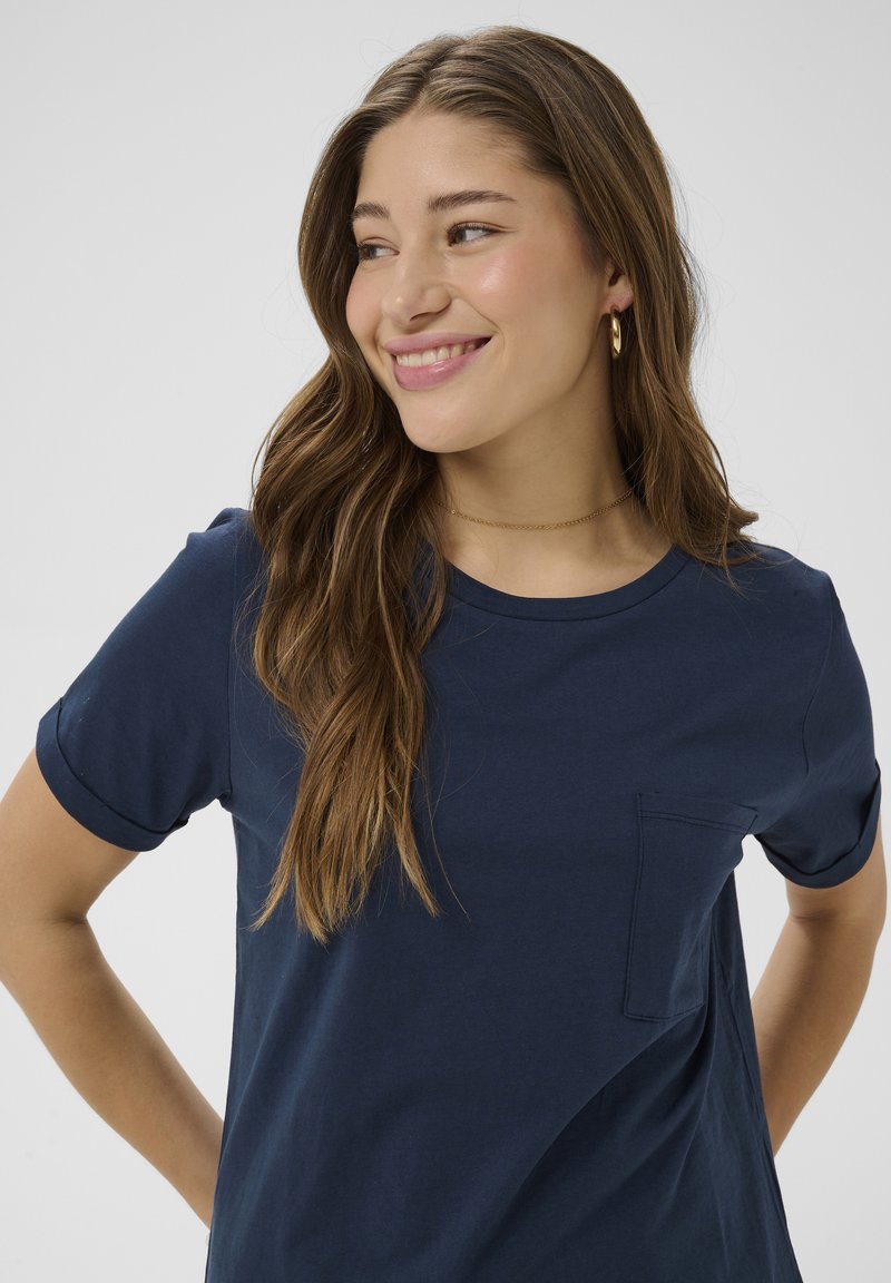 Joven con cabello largo y castaño sonriente, lleva una camiseta azul marino de manga corta con bolsillo frontal y pendientes de aro dorados.