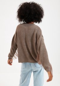 Brązowy sweter dzianinowy o luźnym kroju i długich rękawach, z przezroczystymi panelami po bokach oraz ściągaczami przy mankietach. Widoczne jasnoniebieskie dżinsy.