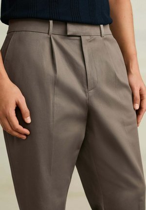 Henkilö, jolla on yllään korkeavyötäröiset taupe-väriset housut, joissa on etuviirut ja vyölenkit, yhdistettynä sisään puettuun mustaan ribbineuletoppaan, kädet rentoina sivuilla.