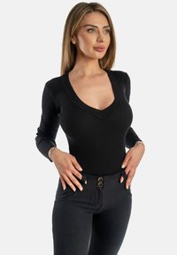 Mustat viiksekset bodysuitit, joissa on V-pääntie ja pitkät hihat, yhdistettynä korkeavyötäröisiin mustiin housuihin, joissa on kultaiset nappikoristeet.