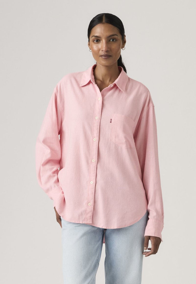 Kvinde med mørkt hår iført en løs, lyserød skjorte med knapper og lyseblå jeans, stående mod en ensfarvet lys baggrund.