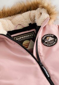Chaqueta rosa claro con un suave forro de forro polar, con borde de capucha de pelaje sintético y cremallera negra. Presenta un parche de logo en el pecho y una etiqueta interna colorida.