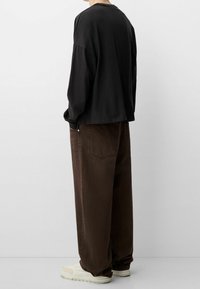 Camisa negra de manga larga con un ajuste relajado, combinada con pantalones marrones holgados y zapatillas crema, con costuras visibles en los pantalones.