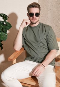 Olivově zelené tričko s kulatým výstřihem vyrobené z měkké látky, spárované s bílými slim fit kalhotami. Model sedí na dřevěné židli s rostlinou v blízkosti.
