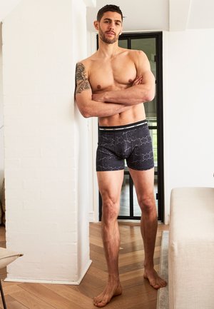 Sorte boxershorts med et marmorert mønster, elastisk talje, og en tætsiddende pasform, båret af en mand der står op ad en væg.