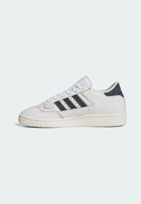 Zapatilla de piel blanca con franjas azul marino, punta redonda, cuello acolchado y suela de goma texturizada. Presenta detalles perforados y cierre de cordones.