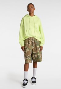 Neon green hoodie with front pocket, paired with brown camo cargo shorts. White socks and black sneakers complete the outfit.