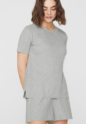 Frau trägt ein hellgraues Oberteil mit kurzen Ärmeln und passende Shorts mit dezenten, wellenförmigen Kanten und steht vor einem schlichten weißen Hintergrund.