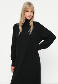 Melnā adīta turtleneck kleita ar brīvu piegriezumu, garām piedurknēm un taisnu siluetu, ar ribotām aprocēm un vidējo garumu.