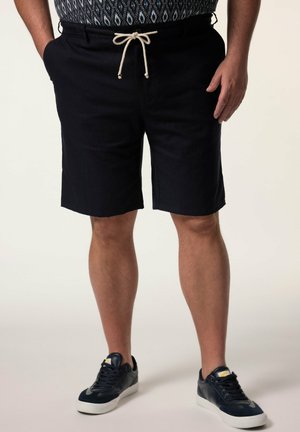 Mand iført sorte knælange shorts med snor, mønstret skjorte stoppet ind, og sorte sneakers med hvide såler mod en ensfarvet baggrund.